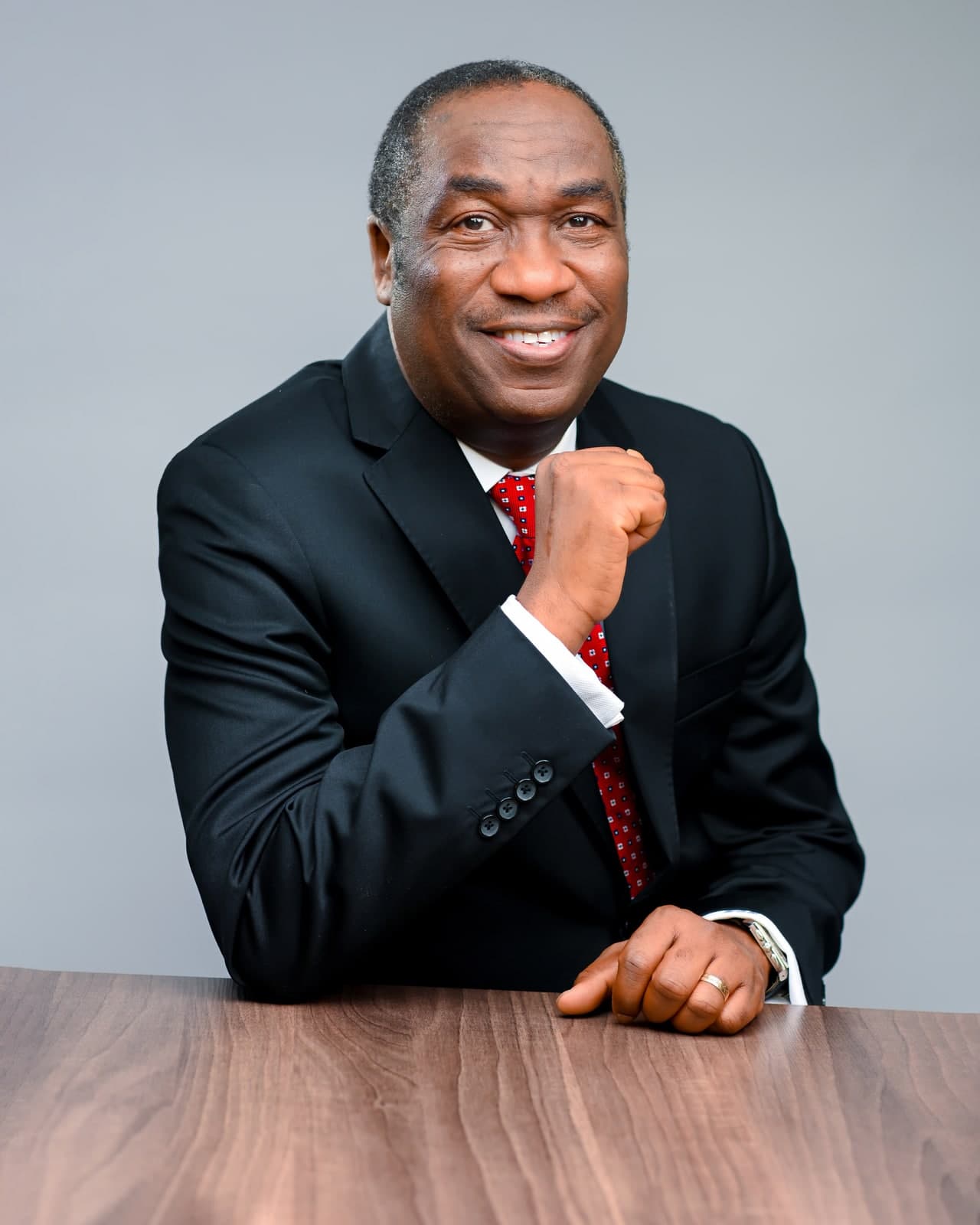 Dr. Kadri Obafemi Hamzat, seated at a wood desk in a dark suit and red tie, fist raised near his chin.