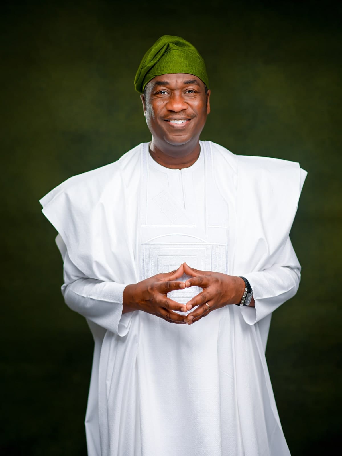 Dr. Kadri Obafemi Hamzat, standing with hands clasped, in a white agbada and green cap, against a dark green backdrop.