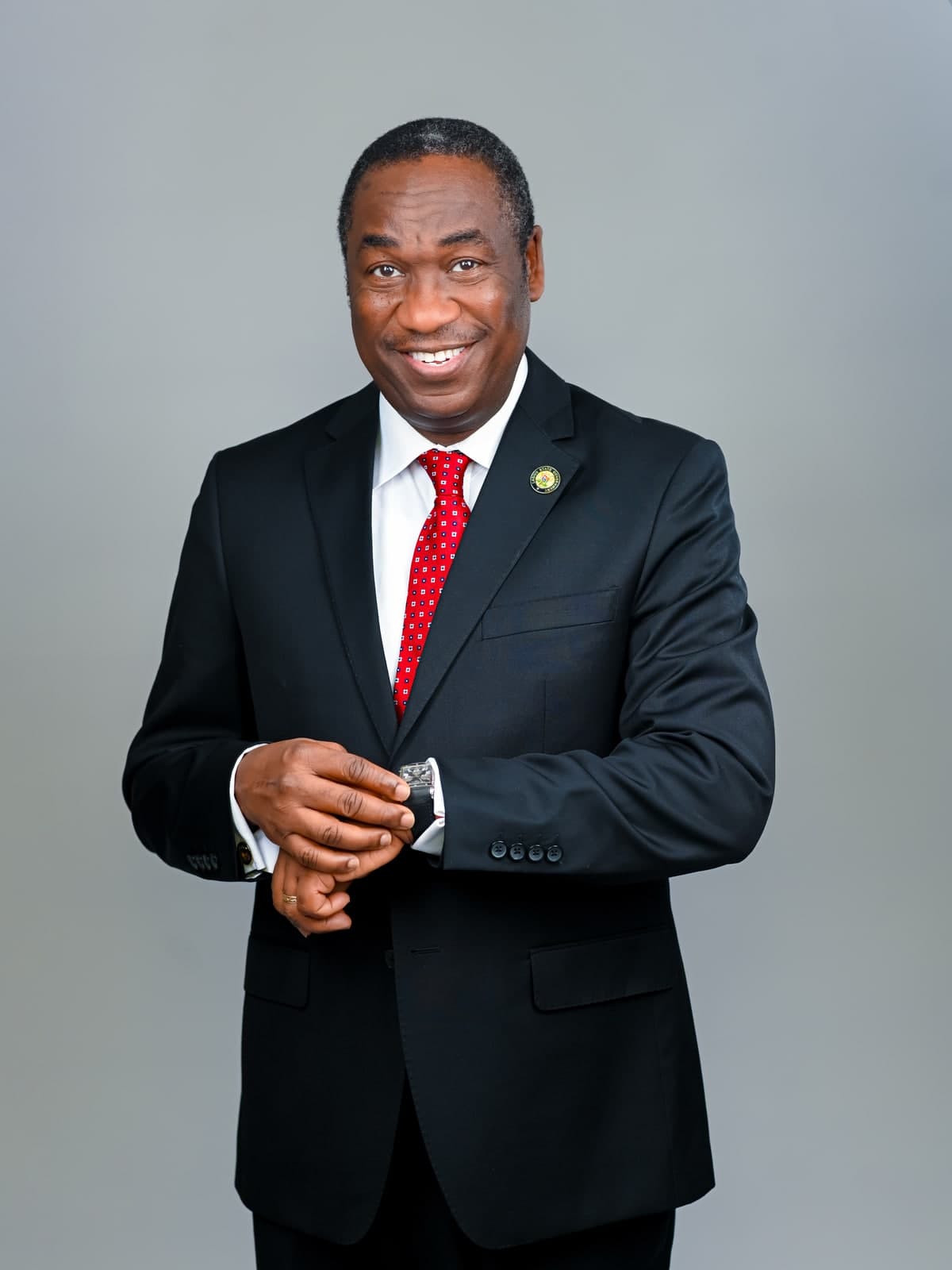 Dr. Kadri Obafemi Hamzat, standing and adjusting his watch, in a dark suit and red tie, against a grey background.
