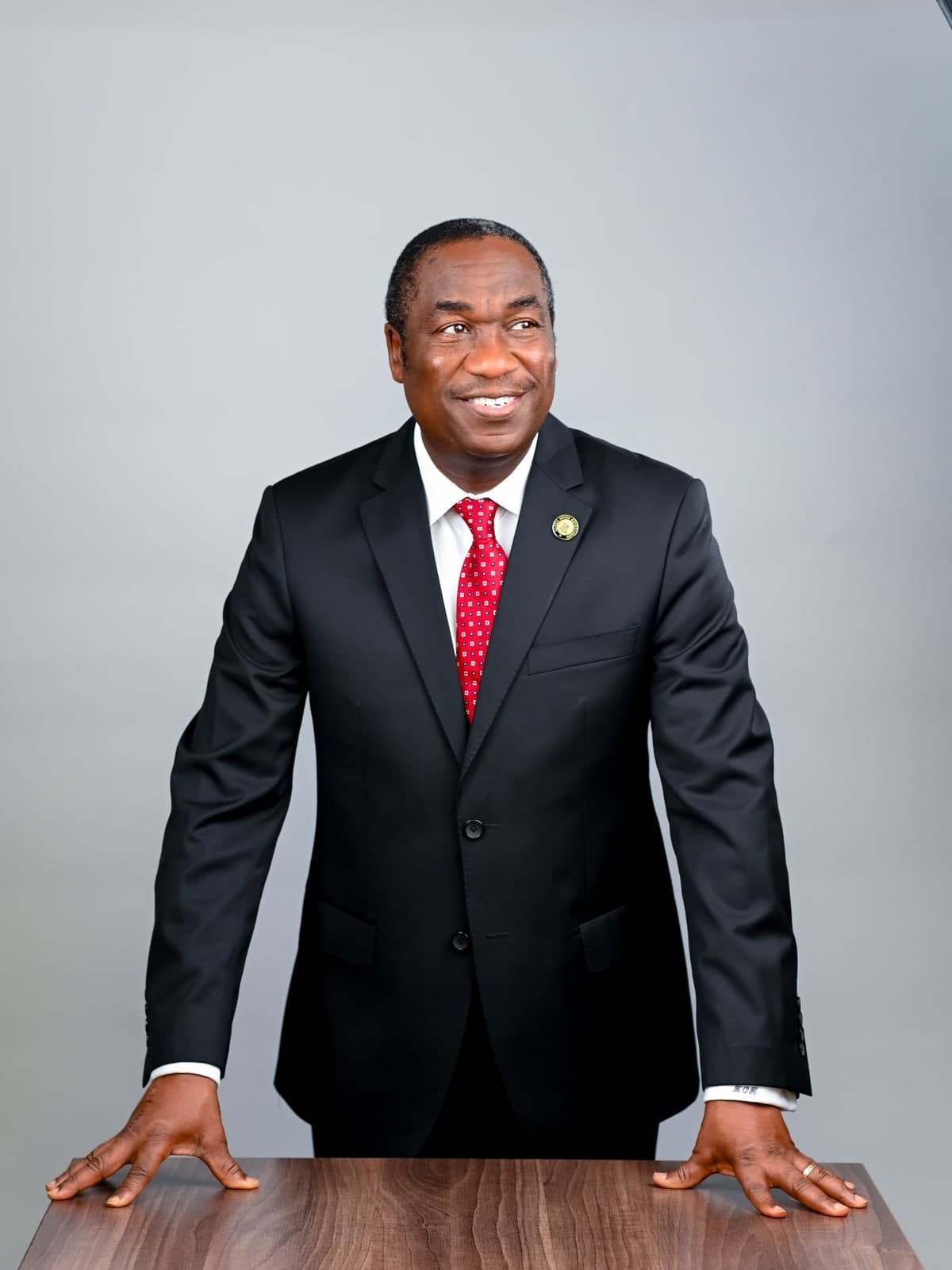 Dr. Kadri Obafemi Hamzat, standing and leaning forward with both hands on a wood table, in a dark suit and red tie.