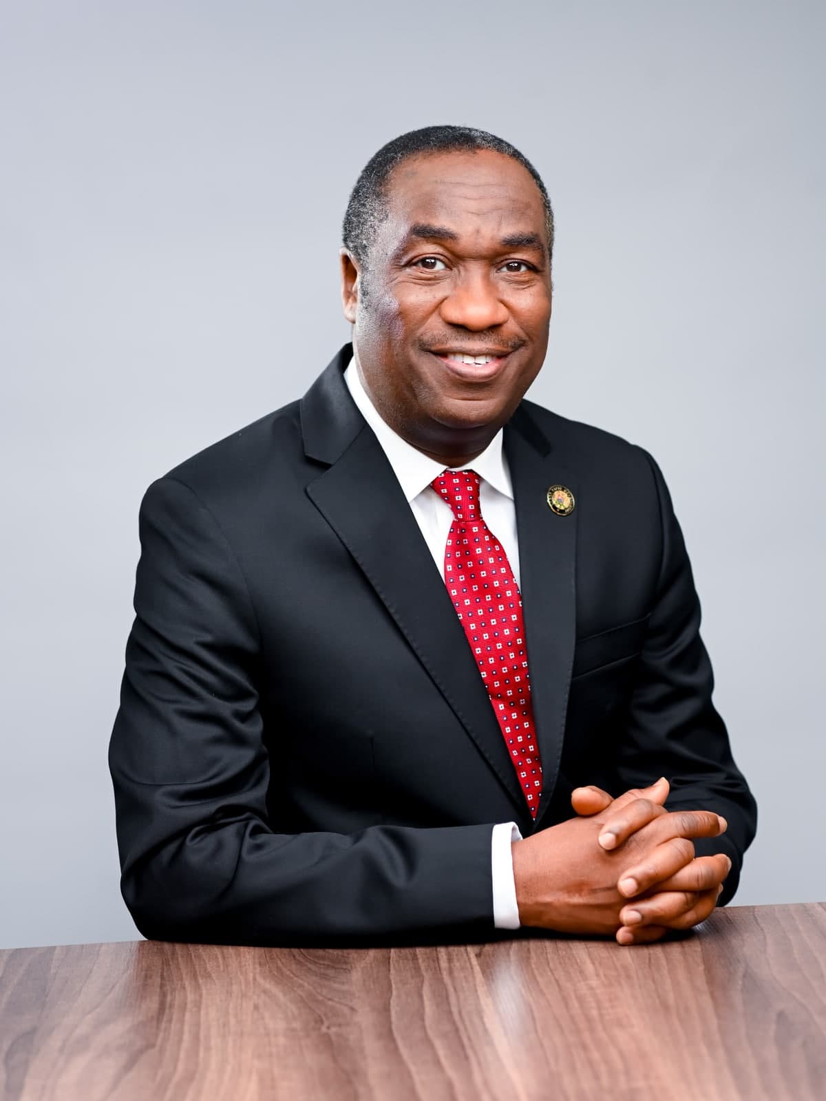 Dr. Kadri Obafemi Hamzat, seated at a wood table with hands folded, in a dark suit and red tie, against a plain grey background.