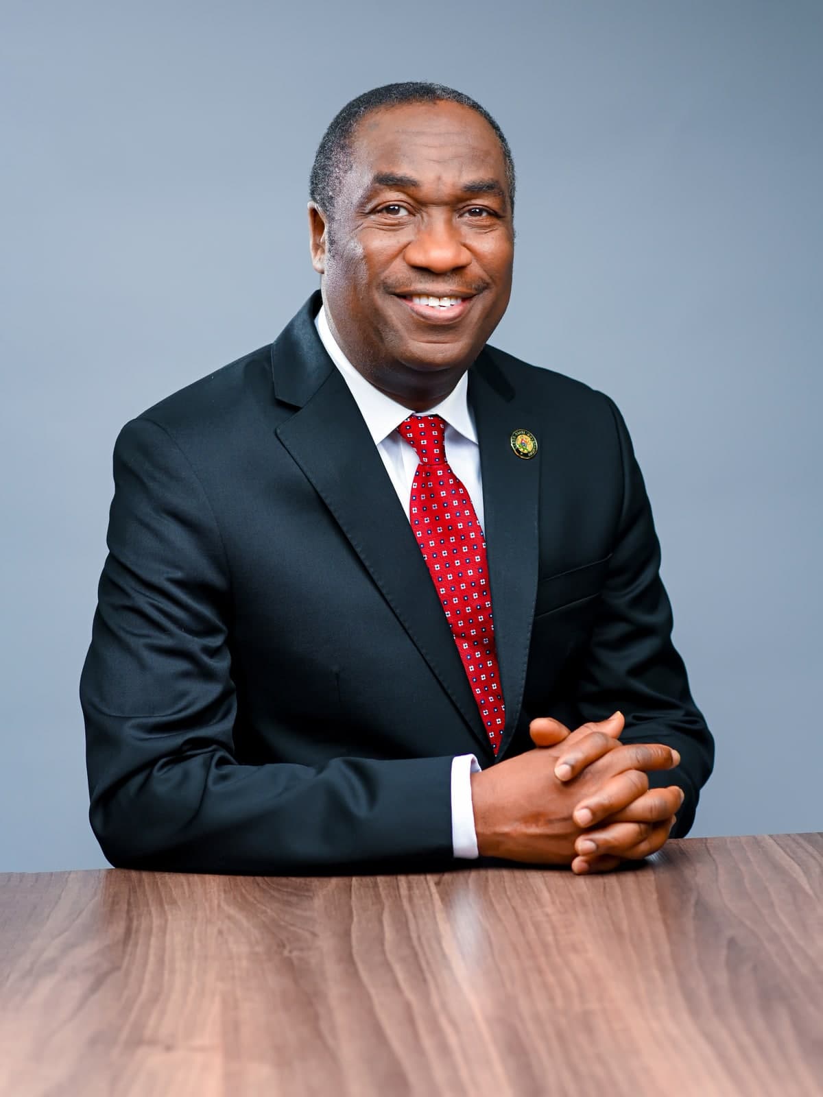Dr. Kadri Obafemi Hamzat, seated at a wood table with hands clasped, smiling directly at the camera, in a dark suit and red tie.