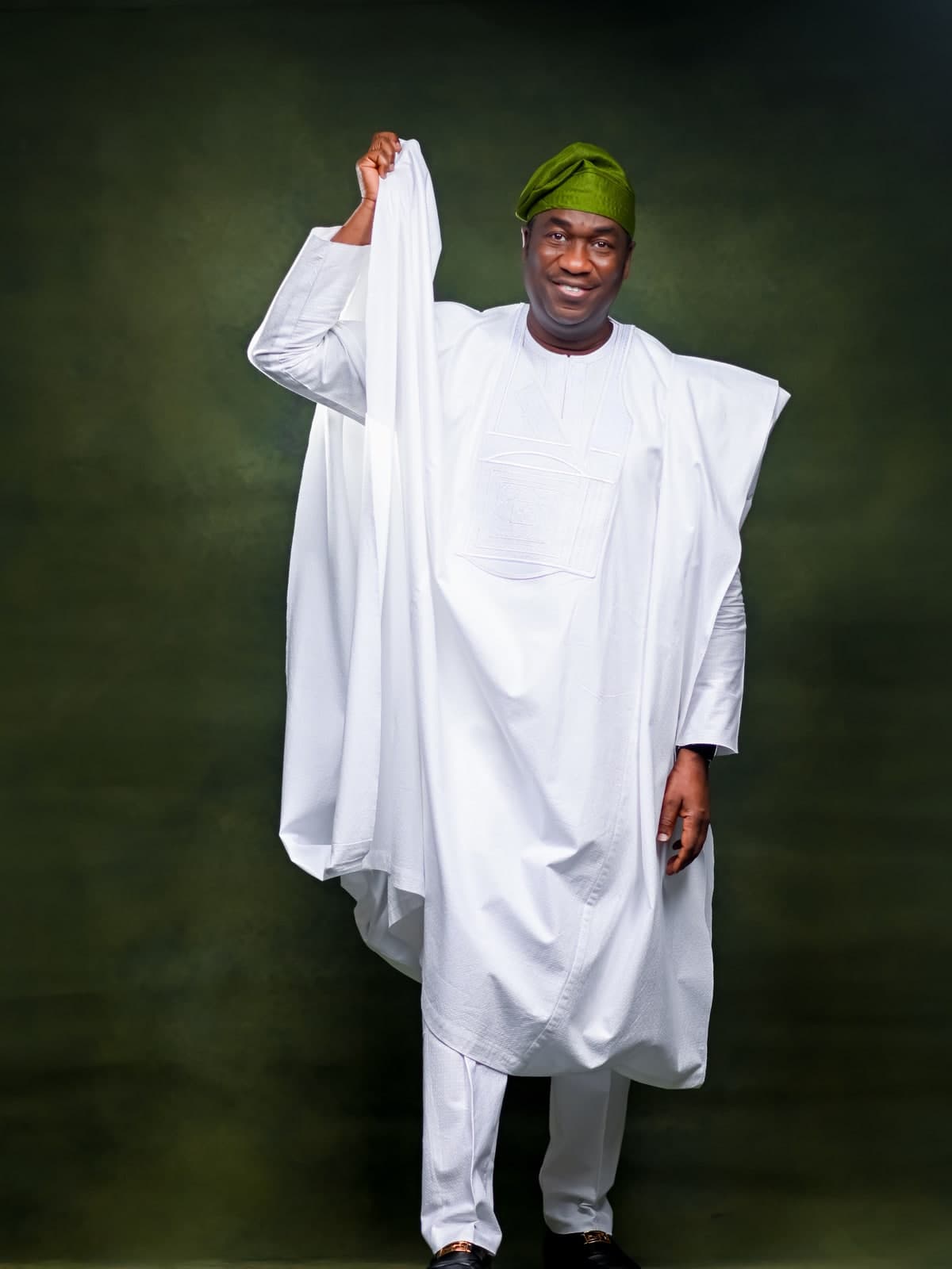 Dr. Kadri Obafemi Hamzat, standing full length in a white agbada and green cap, one hand raised lifting the outer cloth, against a dark green backdrop.