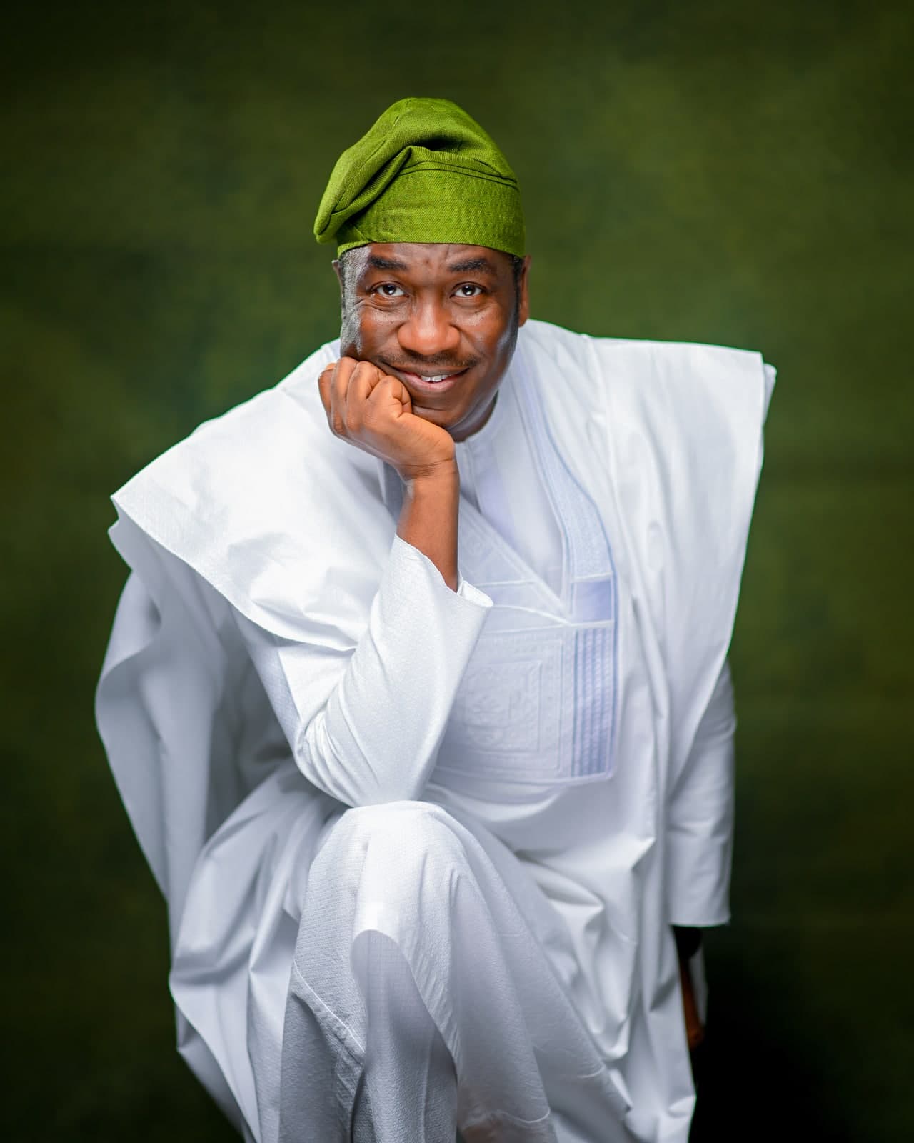 Dr. Kadri Obafemi Hamzat, seated with his chin resting on his fist, in a white agbada and green cap, against a green backdrop.