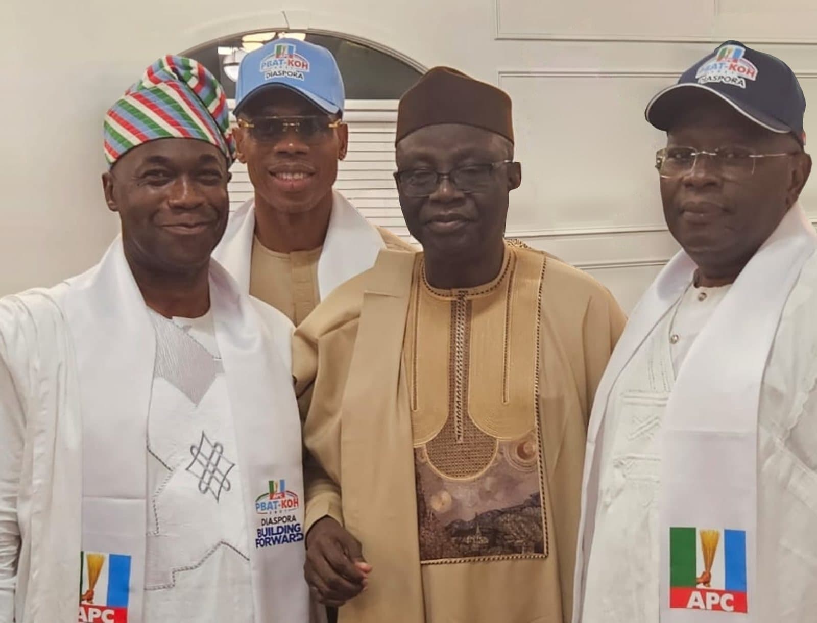 Four men stand together indoors against a white wall: one in a white agbada and a striped cap, one in a light blue PBAT-KOH Diaspora cap and tinted glasses, one in a beige agbada and a brown cap, and one in a navy PBAT-KOH Diaspora cap. Three of them wear white sashes, two with the APC logo, one of which also reads "Building Forward".