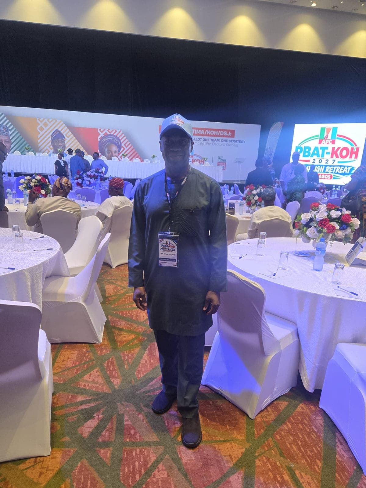 A man in a dark green outfit, a light blue cap and a lanyard stands between round tables with white covers in a banquet hall. A stage with a large screen is behind him.
