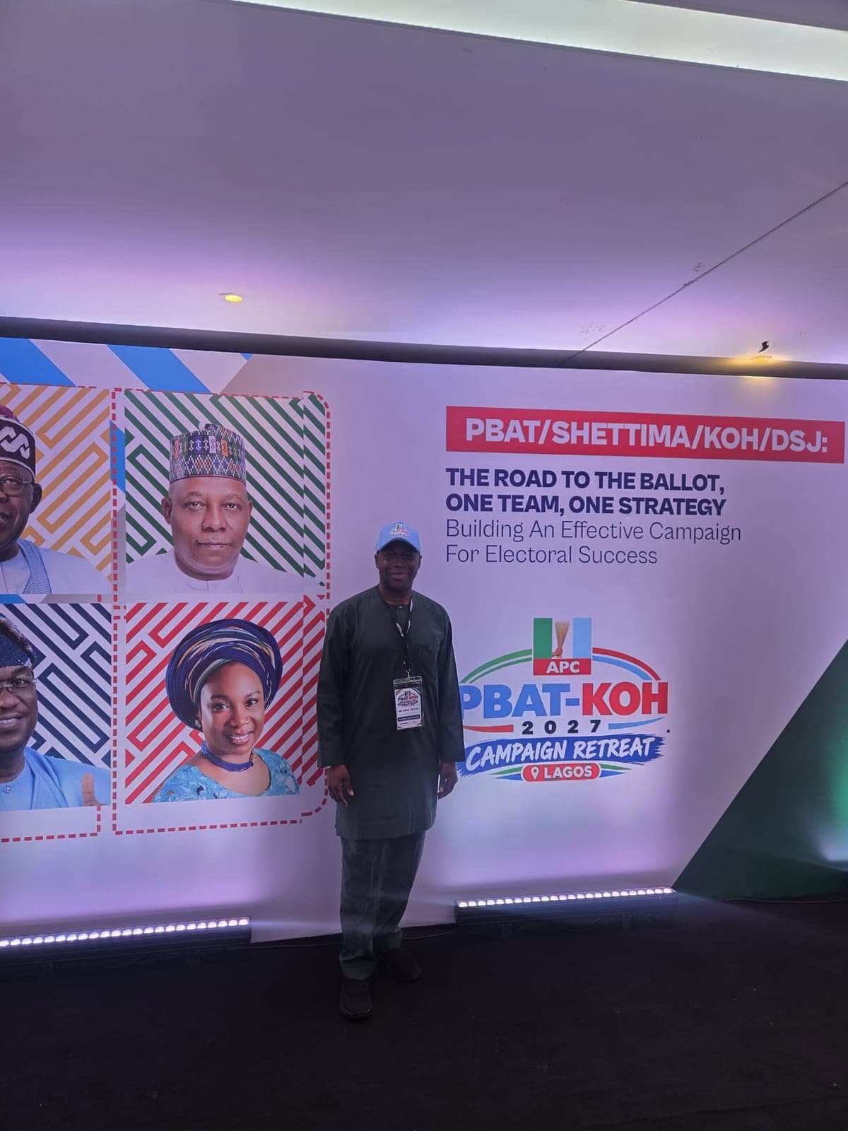 A man in a dark green outfit, a light blue cap and a lanyard stands in front of a backdrop headed "PBAT/SHETTIMA/KOH/DSJ:" with the theme The Road to the Ballot, One Team, One Strategy. The logo reads "APC PBAT-KOH 2027 Campaign Retreat" with "Lagos" beneath it.