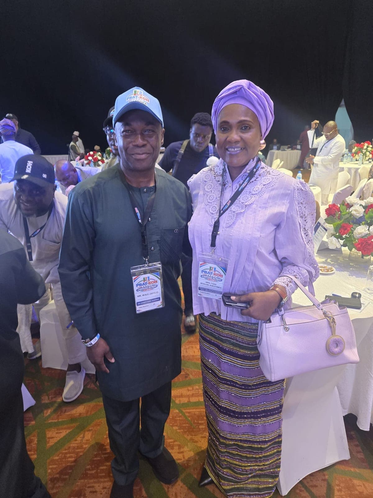 A man in a dark green outfit and a light blue cap and a woman in a lilac head tie, a lilac lace top and a striped skirt, both wearing lanyards, stand together in a banquet hall. The woman holds a white handbag. Round tables with red and white flowers are behind them.