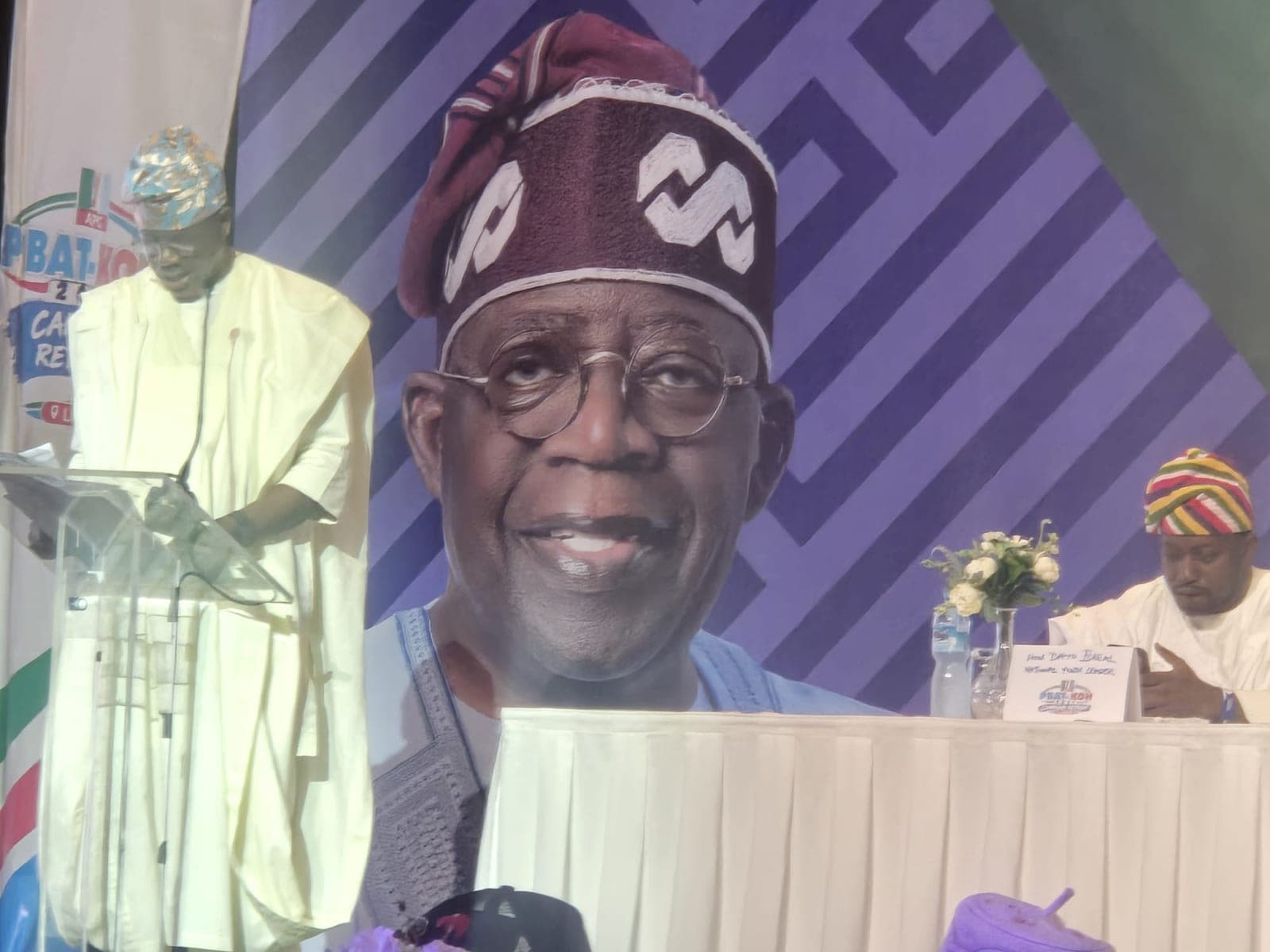 A large screen behind a lectern shows a smiling man in a maroon patterned cap and round glasses. A man in a white robe stands at the lectern, and another man sits at a table to the right with a vase of flowers in front of him.
