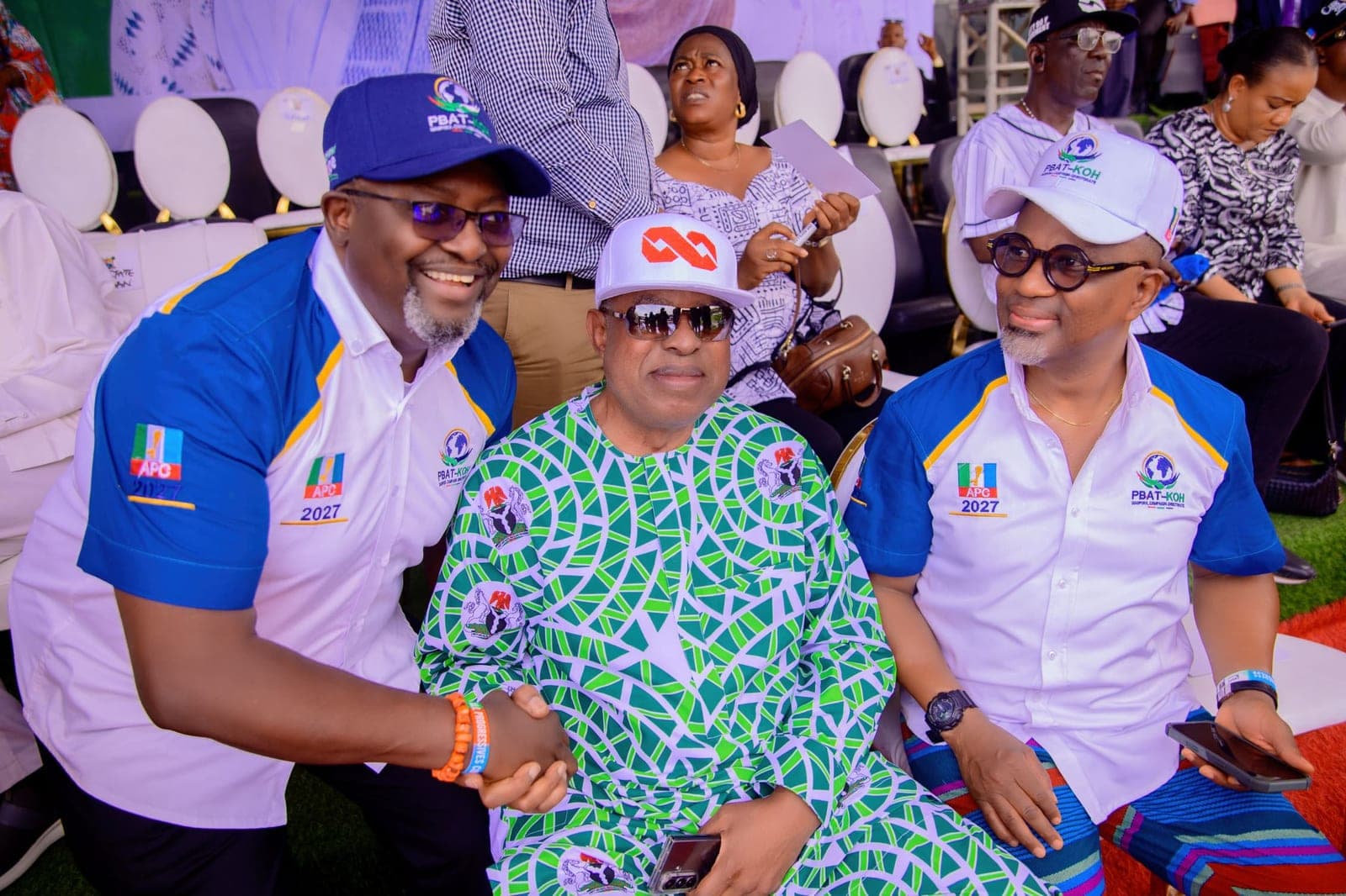 Three men sit close together in an open-air seating area. Two wear white and blue campaign shirts, and between them a man in a green and white patterned shirt wears a white cap and sunglasses. Rows of white chairs are behind them.