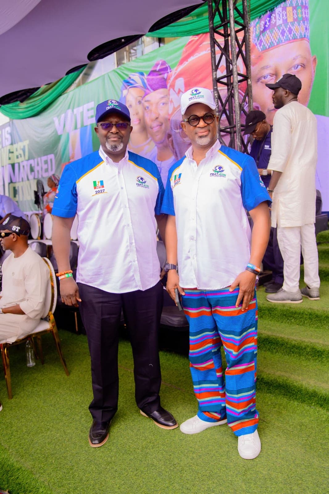 Two men in white and blue campaign shirts and caps stand side by side on green turf in front of a large campaign backdrop. One wears trousers striped in several colours.