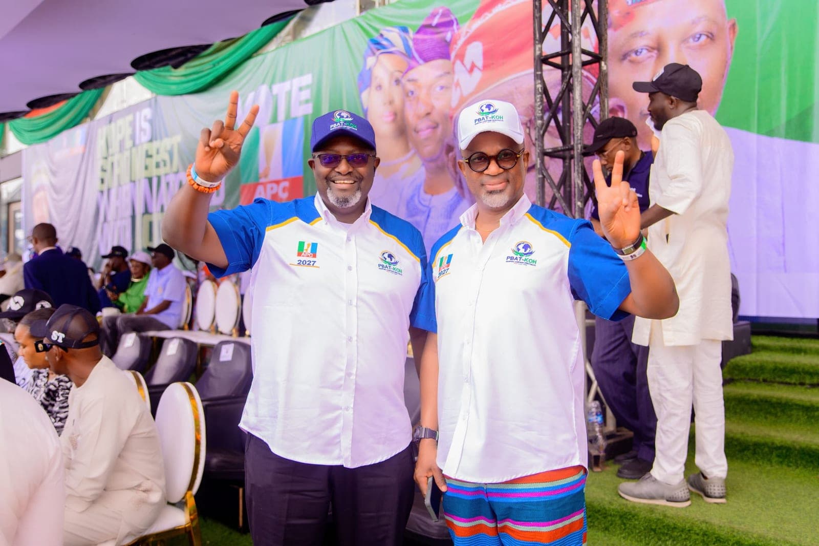 Two men in white and blue campaign shirts and caps stand side by side, each raising a hand with two fingers up, in front of a backdrop with large lettering ending "is strongest".