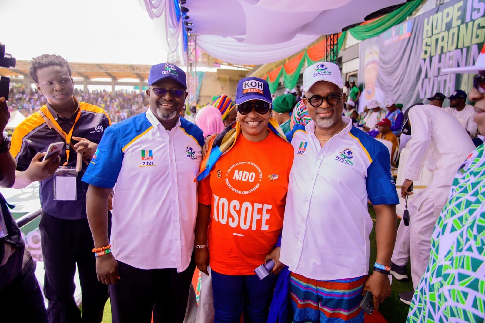 Two men in white and blue campaign shirts and caps pose with a woman in an orange T-shirt and a cap between them, at an outdoor event with other people and draped canopies behind them.