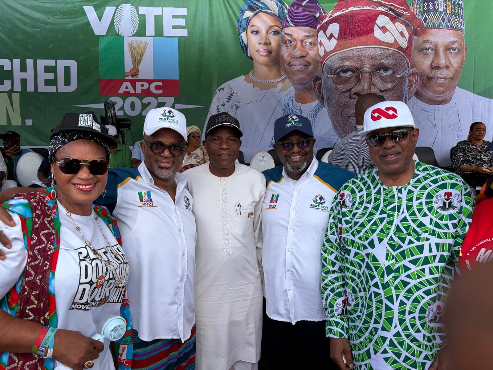Five people stand in a row in front of a large green backdrop that reads "Vote APC 2027", with portraits on the backdrop above them. From left: a woman in a patterned shawl, a black cap and sunglasses; two men in white and blue campaign shirts and caps; a man in a white robe; and a man in a green and white patterned shirt and a white cap. The caption it was shared with names three people and does not say which is which.