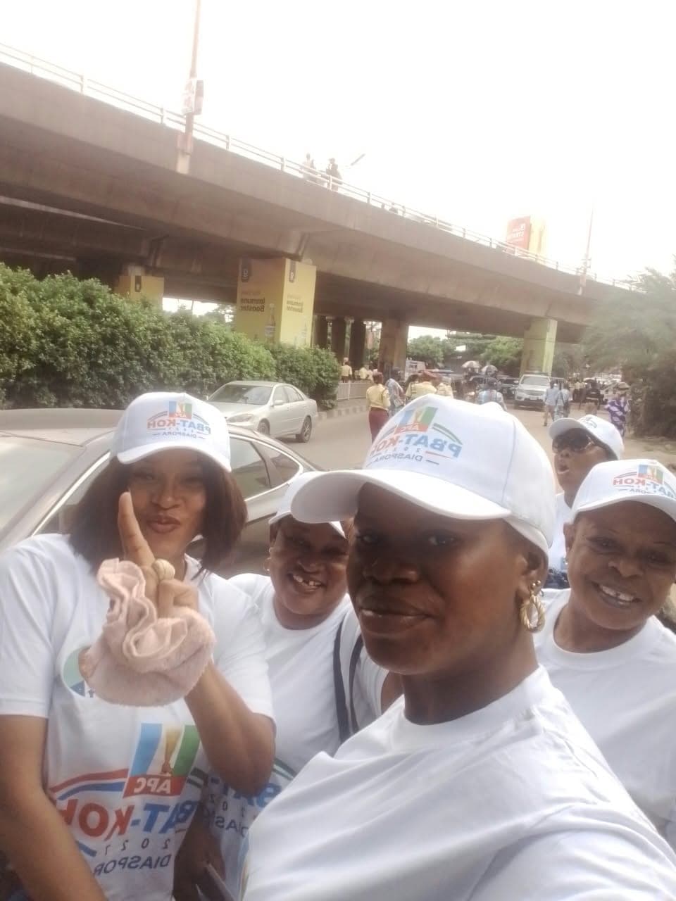 Women in white campaign caps and white T-shirts stand close together outdoors in front of a road overpass. One of them holds up two fingers.
